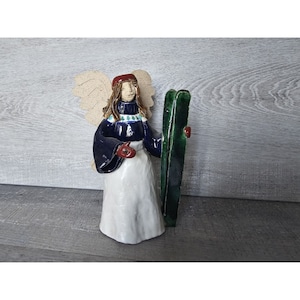 Puede incluir: Una figurita de ángel de cerámica con alas, que lleva un suéter azul con un patrón blanco y verde, un gorro rojo y sostiene un esquí verde.