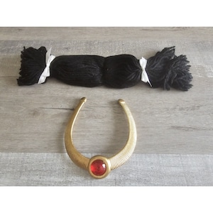 May include: A gold-colored necklace with a red jewel in the center. The necklace is on a wooden surface. There is a bundle of black yarn with white ribbons on the surface as well.