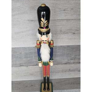 Puede incluir: Un cascanueces de madera con un sombrero negro, adornos dorados y una barba blanca. El cascanueces lleva un uniforme azul y verde con pantalones rojos y botas negras. El cascanueces está de pie sobre una base verde.