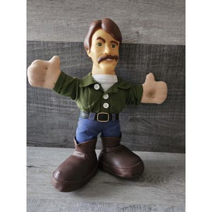 May include: A plush doll of a man with brown hair, a mustache, and a green shirt. He is wearing blue jeans and brown boots. He is giving a thumbs up with both hands.