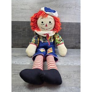 May include: A vintage rag doll with red yarn hair, a painted face, and a blue and white hat. The doll wears a floral print shirt, blue overalls, and red and white striped legs. Black shoes complete the outfit.