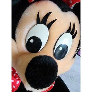 Pu&ograve; includere: Un giocattolo di peluche di un personaggio dei cartoni animati con grandi occhi neri, un naso nero e un fiocco rosso con pois bianchi. Il personaggio ha un viso marrone chiaro e ciglia nere.