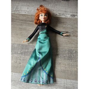 May include: A red-haired doll wearing a black and green dress with gold accents. The doll has a long green skirt with a peacock design at the bottom.