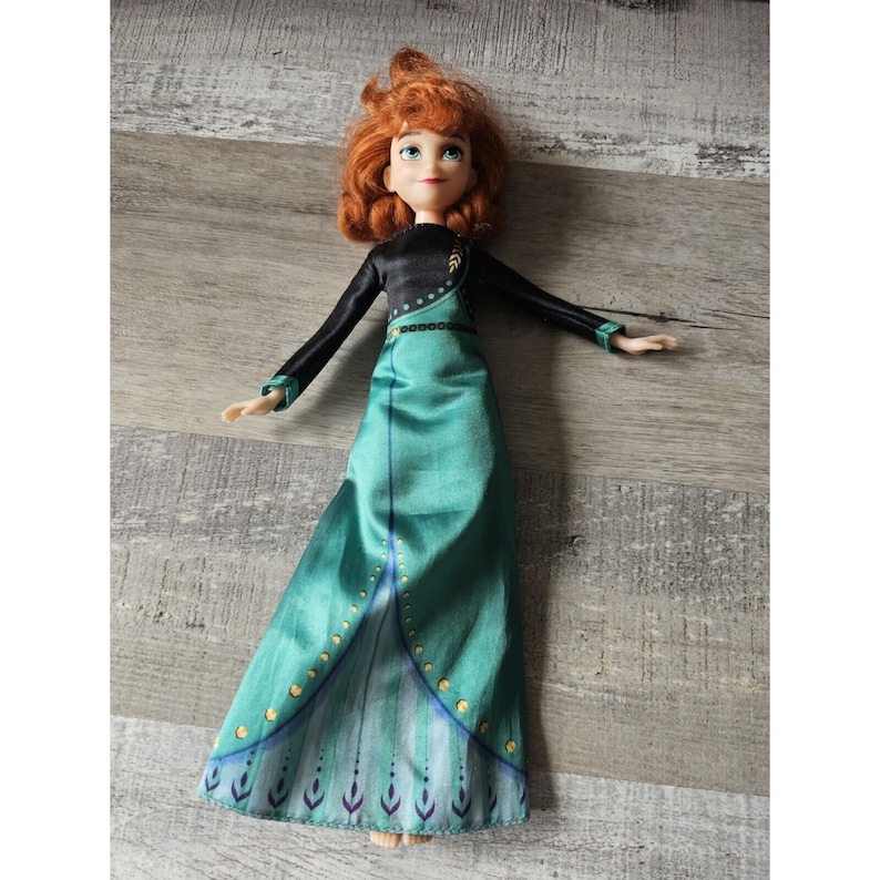 May include: A red-haired doll wearing a black and green dress with gold accents. The doll has a long green skirt with a peacock design at the bottom.
