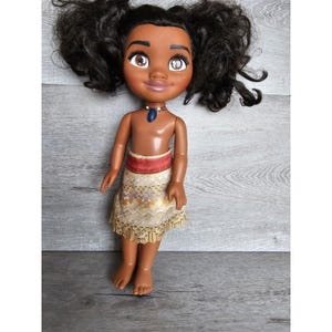May include: A brown-skinned doll with black curly hair, wearing a gold and brown skirt with a red sash and a blue necklace. The doll has large brown eyes and a friendly expression.