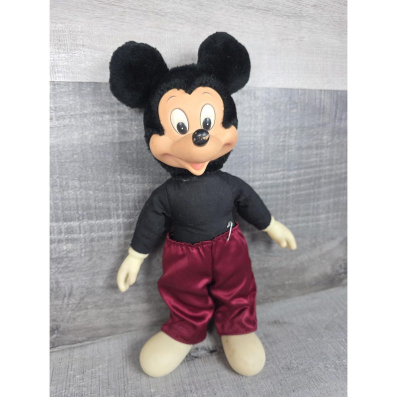 May include: Vintage Mickey Mouse plush doll. The doll has a black shirt, burgundy pants, and large black ears. The face is tan with black eyes and a black nose. The hands and feet are cream-colored.