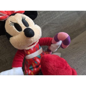 May include: A plush Minnie Mouse doll wearing a red dress with a white belt and a red bow. The doll is holding a candy cane with pink, purple, and white stripes. The doll has black ears and a black nose.