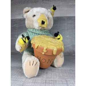 Pode incluir: Um urso de pelúcia vestindo um suéter verde, coberto de mel amarelo e segurando um vaso de terracota cheio de mel. O vaso tem a palavra "HON" escrita. Duas pequenas figuras de abelha estão presas ao urso.