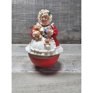 May include: A ceramic Mrs. Claus figurine in a red dress and white apron, holding a teddy bear and a doll. The figurine has a round red base and a white curly wig. A small teddy bear is in her apron pocket.
