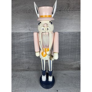 Puede incluir: Una figura de cascanueces de madera con forma de conejo que lleva un sombrero de copa rosa pálido con orejas largas. La figura tiene una barba blanca, un chaleco amarillo y pantalones a rayas azules y blancas. Un pequeño reloj de bolsillo dorado cuelga.