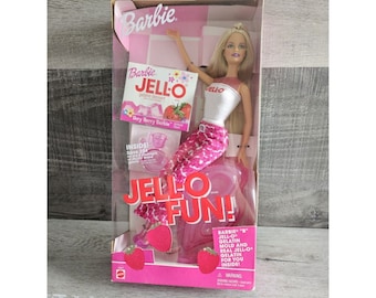 May include: A pink and white Barbie doll wearing a white tank top and pink and white patterned trousers. The doll is sitting on a pink and white Jell-O mould. The box also includes a packet of Jell-O gelatin mix and a pink jug. The box reads "Barbie "B" Jell-O Gelatin Mould and Real Jell-O Gelatin for You Inside!"