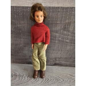 May include: A vintage doll with brown hair, wearing a red turtleneck sweater and green pants. The doll is wearing brown shoes.