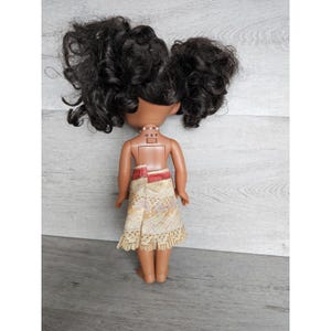 May include: A brown-skinned doll with long black curly hair styled in two pigtails. The doll is wearing a yellow and brown patterned skirt with a red waistband and a white necklace with a small charm. The doll's back is visible, and the text "OXT" is printed on the doll's back.