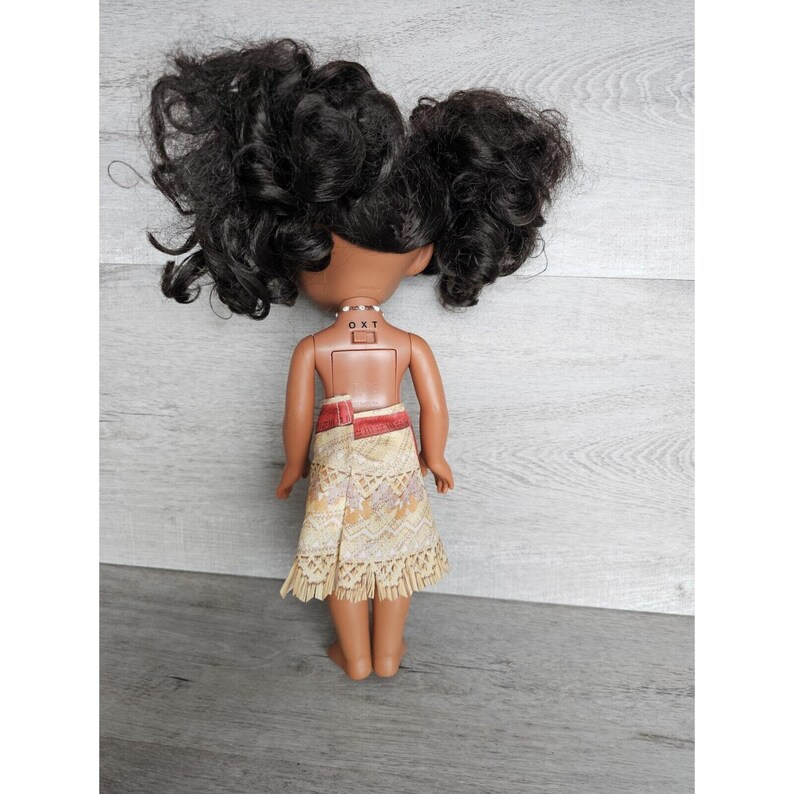 May include: A brown-skinned doll with long black curly hair styled in two pigtails. The doll is wearing a yellow and brown patterned skirt with a red waistband and a white necklace with a small charm. The doll's back is visible, and the text "OXT" is printed on the doll's back.