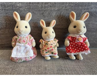 May include: Three toy rabbits wearing dresses. The rabbit on the left has a pink floral dress and a white apron. The middle rabbit has a yellow floral dress. The rabbit on the right has a red polka dot dress.
