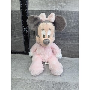 Puede incluir: Un mu&ntilde;eco de peluche de Minnie Mouse sentado. El mu&ntilde;eco tiene orejas grises y un lazo rosa. El cuerpo es rosa con detalles blancos en las manos y los pies. La cara es rosa claro con pesta&ntilde;as negras y nariz gris.