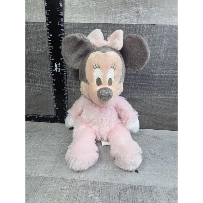 Puede incluir: Un mu&ntilde;eco de peluche de Minnie Mouse sentado. El mu&ntilde;eco tiene orejas grises y un lazo rosa. El cuerpo es rosa con detalles blancos en las manos y los pies. La cara es rosa claro con pesta&ntilde;as negras y nariz gris.