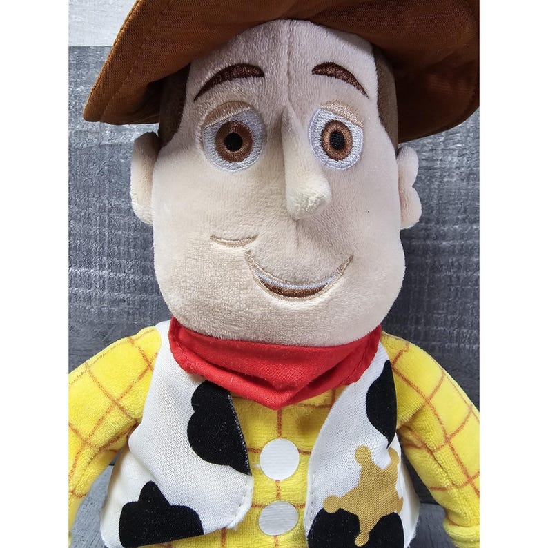 May include: A plush toy of a cowboy character with a brown hat, a yellow plaid shirt, and a white vest with black cow spots. The character has a red neckerchief and a sheriff's badge.