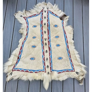 May include: Hand-painted hide rug with a cream background and a decorative border. The border features a blue and maroon scalloped design with oval blue accents. The rug has a fur trim.