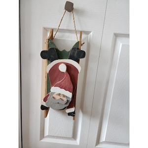 May include: A wooden Santa Claus ornament hanging from a door. Santa is painted red, white, and grey and is holding onto a green and brown wooden sledge.