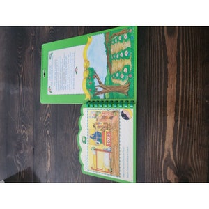 May include: A green and yellow children's book with a spiral binding. The front cover features a cartoon illustration of a toy store with the word "TOYS" above the door. The back cover features a cartoon illustration of a tree with apples and a row of children's faces peeking out from behind a green hedge. The book title is "Baby's Favorite Bedtime Story" and the subtitle is "Baby's First Bedtime Snack".