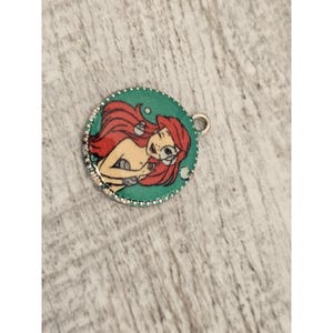May include: A round, silver charm with a green background and a picture of Ariel, the Little Mermaid, with red hair and a seashell in her hand. There are white bubbles around her.
