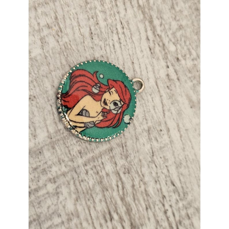 May include: A round, silver charm with a green background and a picture of Ariel, the Little Mermaid, with red hair and a seashell in her hand. There are white bubbles around her.