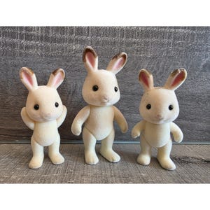 May include: Three cream-colored rabbit figurines with brown-tipped ears and black eyes stand in a row. The rabbits have a soft, flocked texture and are posed in different positions, giving them a playful appearance.