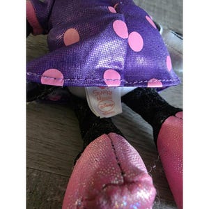 Op de afbeelding: Een close-up van een Minnie Mouse knuffel. Het speelgoed heeft een paarse jurk met roze stippen, roze schoenen en zwarte benen. Het label luidt "Sparkle Minnie" en "Disney".