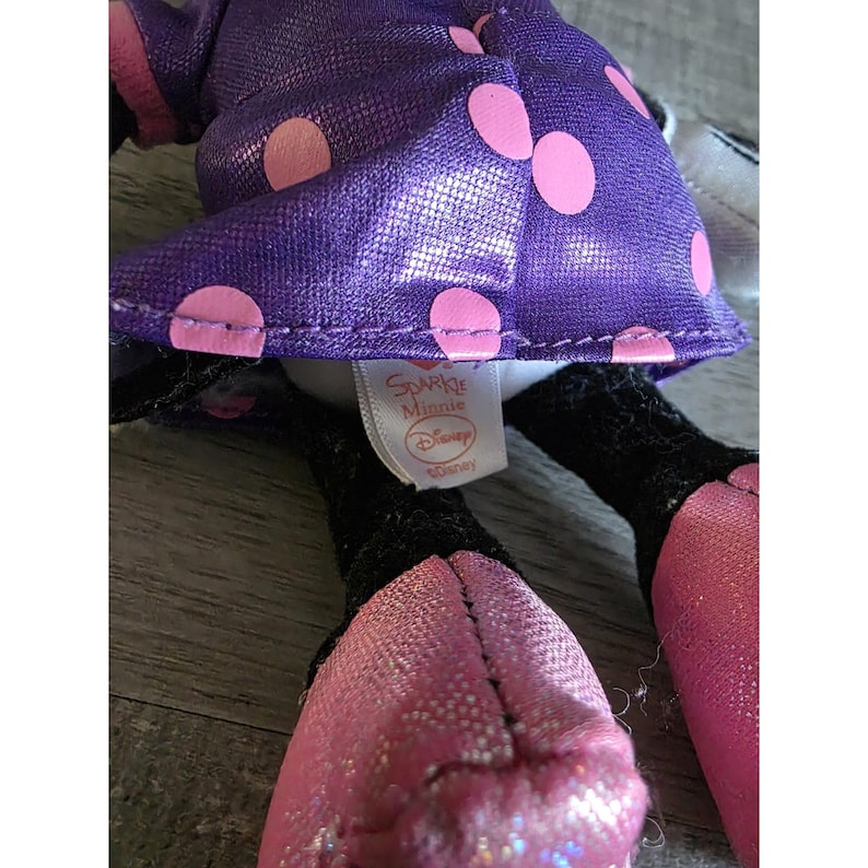 Op de afbeelding: Een close-up van een Minnie Mouse knuffel. Het speelgoed heeft een paarse jurk met roze stippen, roze schoenen en zwarte benen. Het label luidt "Sparkle Minnie" en "Disney".