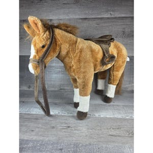 Puede incluir: Un caballo de peluche marrón con marcas blancas en la cara y las patas. Cuenta con una silla de montar y una brida marrones. El caballo de juguete está sobre una superficie de madera.