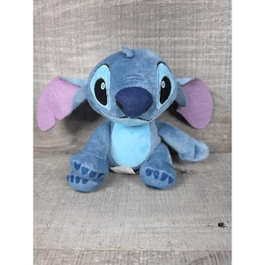 May include: A plush toy character featuring soft blue fur, a large dark blue nose, and oversized ears with light purple inner sections. The character has large black eyes with white highlights, a light blue belly patch, and stitched paw details.