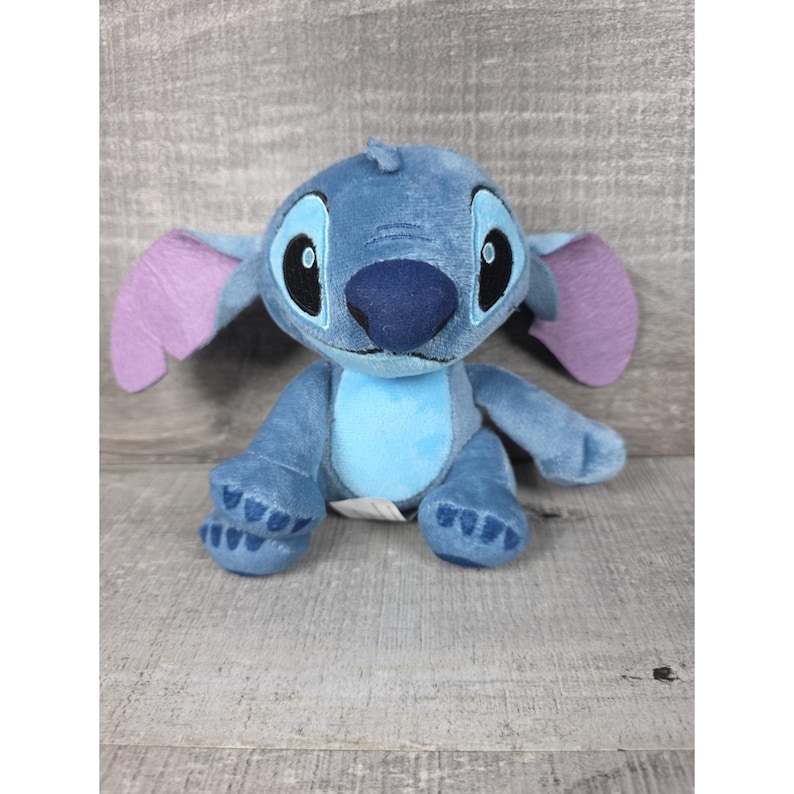 May include: A plush toy character featuring soft blue fur, a large dark blue nose, and oversized ears with light purple inner sections. The character has large black eyes with white highlights, a light blue belly patch, and stitched paw details.