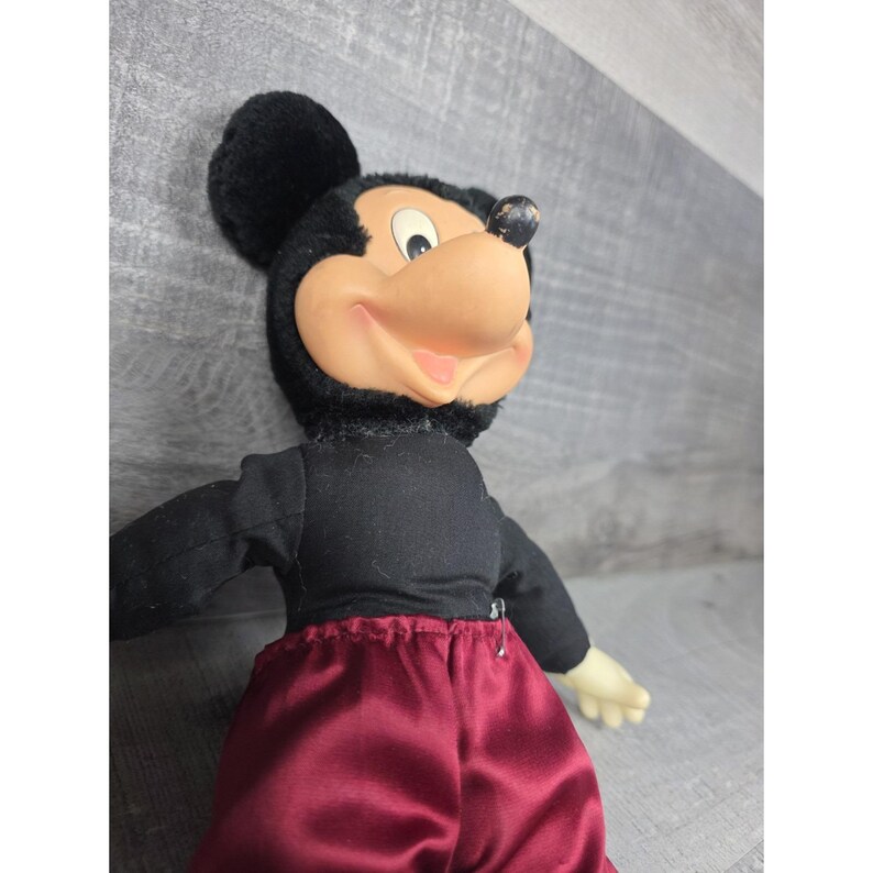 May include: A vintage Mickey Mouse doll with a black body and head, a tan face, and red satin pants. The doll has black ears and a smiling expression. The doll's arms are outstretched, and the background is a light gray.