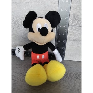 Puede incluir: Un juguete de peluche de Mickey Mouse con orejas negras, cuerpo negro, pantalones cortos rojos con dos botones blancos y zapatos amarillos. El juguete est&aacute; sentado sobre una superficie de madera.