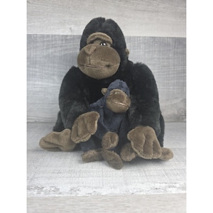 May include: A plush gorilla and baby gorilla toy. The adult gorilla is black with brown accents on the face, hands, and feet. The baby gorilla is dark blue with brown accents. Both are sitting on a light gray wooden surface.