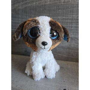 May include: A plush dog toy with a white body, brown and white head, and large blue eyes. The dog has brown ears with blue accents and is sitting upright. The toy is designed to resemble a puppy.