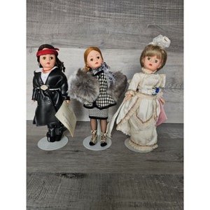 May include: Three porcelain dolls dressed in different outfits. The doll on the left is wearing a black leather jacket, a red bandana, and black pants. The doll in the middle is wearing a black and white checkered dress with a fur collar and black boots. The doll on the right is wearing a white dress with lace trim and a white hat.