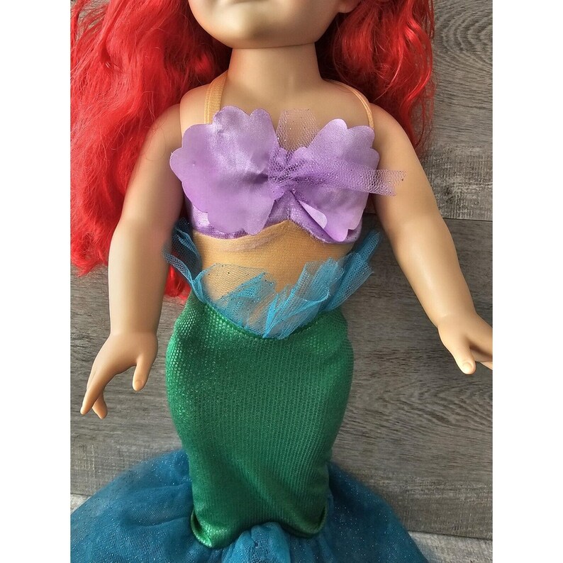 Puede incluir: Una mu&ntilde;eca de pelo rojo con un disfraz de sirena morado y dorado. El disfraz tiene un top morado de concha de mar con una falda de tul dorada y una cola de sirena verde.