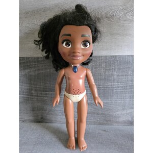 May include: A doll with dark skin, black curly hair, and large brown eyes. The doll wears a white diaper and a blue pendant necklace. The doll's torso has a speaker. The doll is standing on a gray surface.