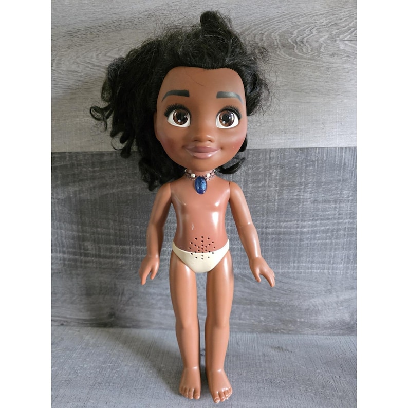 May include: A doll with dark skin, black curly hair, and large brown eyes. The doll wears a white diaper and a blue pendant necklace. The doll's torso has a speaker. The doll is standing on a gray surface.