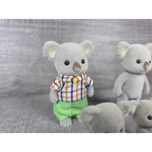 May include: Several gray koala figurines. One koala wears a plaid shirt with orange, blue, and white squares, green pants, and a yellow button. Other koalas are visible in the background.