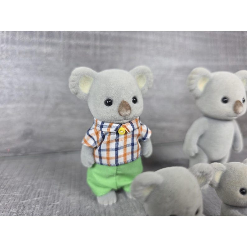 May include: Several gray koala figurines. One koala wears a plaid shirt with orange, blue, and white squares, green pants, and a yellow button. Other koalas are visible in the background.