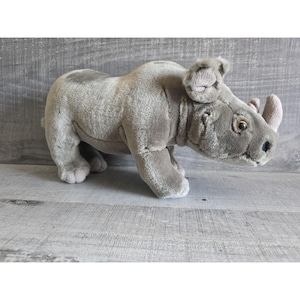 Pode incluir: Um brinquedo de rinoceronte de pelúcia cinza com um chifre cinza claro e orelhas pequenas e detalhadas. O animal de pelúcia está em pé sobre as quatro patas, com um design realista. O brinquedo é feito de material macio e felpudo.