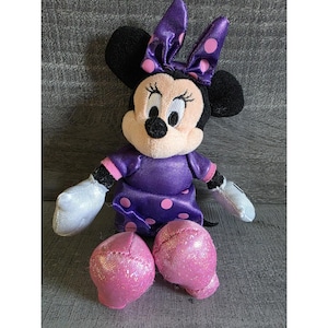 Op de afbeelding: Een zittende Minnie Mouse pluche pop. De pop heeft zwarte oren, een lichtroze gezicht en een paarse jurk met roze stippen. De strik op haar hoofd en haar schoenen zijn ook paars met roze stippen. Haar handschoenen zijn wit.