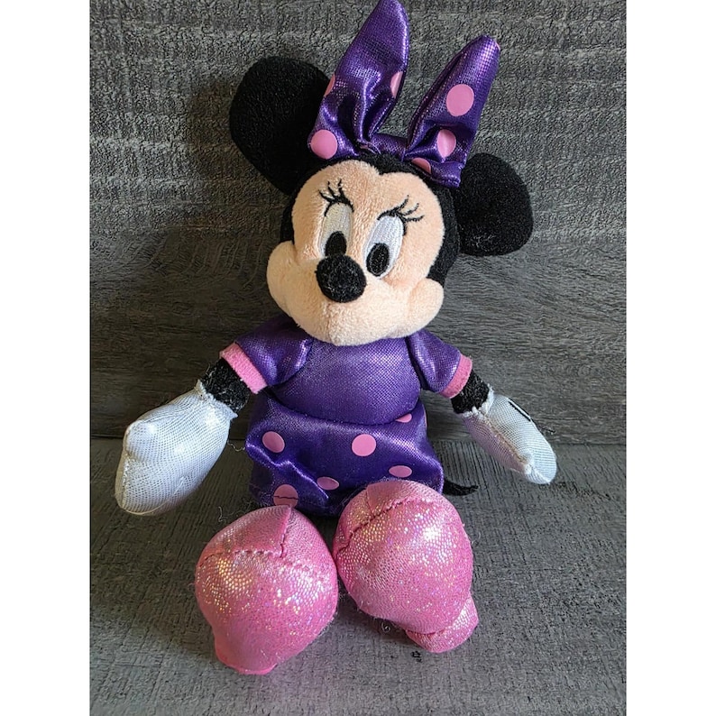 Op de afbeelding: Een zittende Minnie Mouse pluche pop. De pop heeft zwarte oren, een lichtroze gezicht en een paarse jurk met roze stippen. De strik op haar hoofd en haar schoenen zijn ook paars met roze stippen. Haar handschoenen zijn wit.