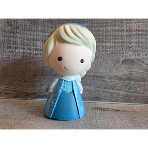May include: A figurine of a cartoon character with a light skin tone, blonde hair, and a blue dress. The figurine has a smiling expression and is standing upright. The dress has a detailed design. The figurine is likely a collectible item.