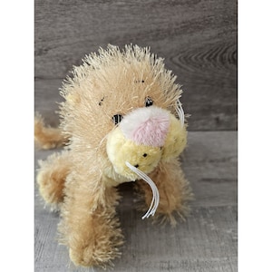 May include: A plush brown lion with a pink nose and yellow fur around its mouth. The lion has white whiskers and is sitting on a wooden surface.