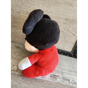 以下が含まれることがあります： 赤と白の服を着たミッキーマウスのぬいぐるみ。おもちゃは木製の表面に座っています。