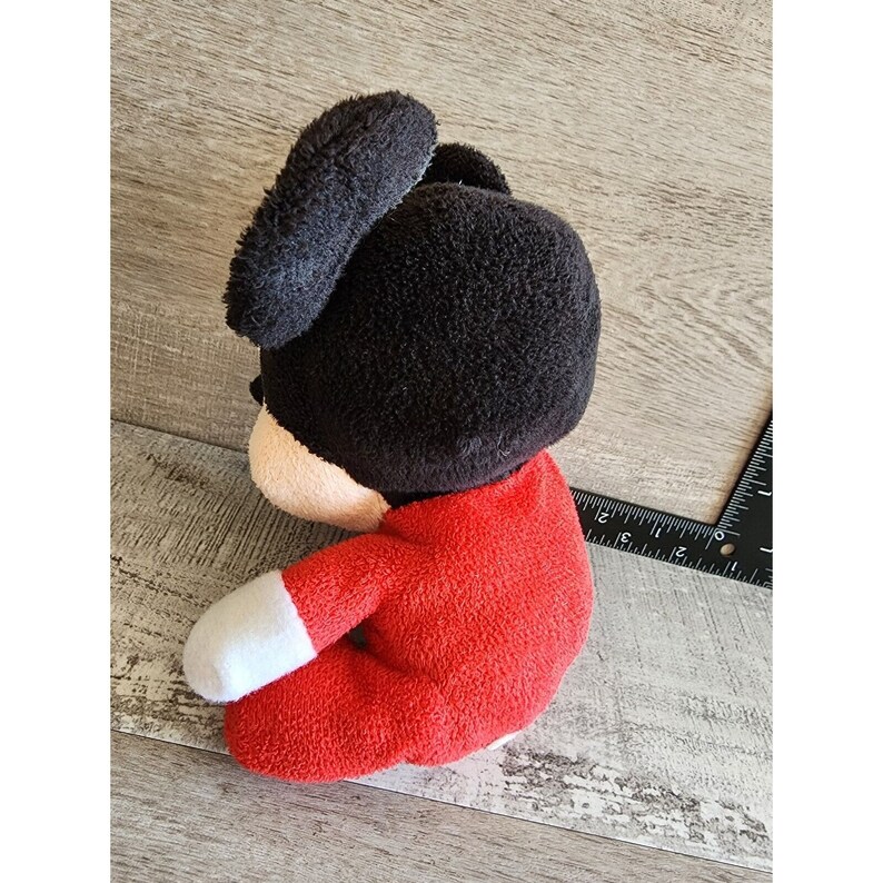 以下が含まれることがあります： 赤と白の服を着たミッキーマウスのぬいぐるみ。おもちゃは木製の表面に座っています。